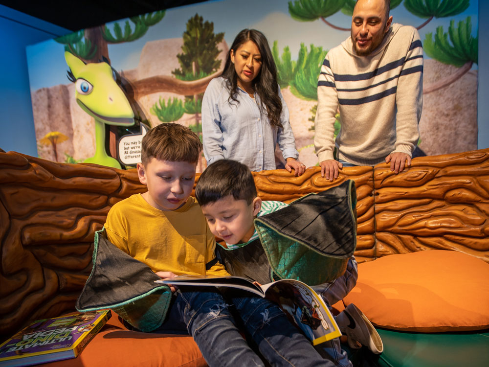 Two children sitting in a giant nest looking at a book Two adults and Mrs. Pteranodon cutout are looking over them from behind.